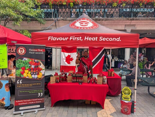 Kiosk set outdoors at a community market featuring person showing off a bottle of hot sauce.