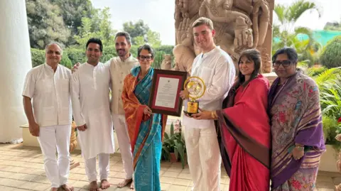 Sebbie Hall Kindness Foundation A group of people in south Asian attire are smiling at the camera while the two people in the middle are holding awards